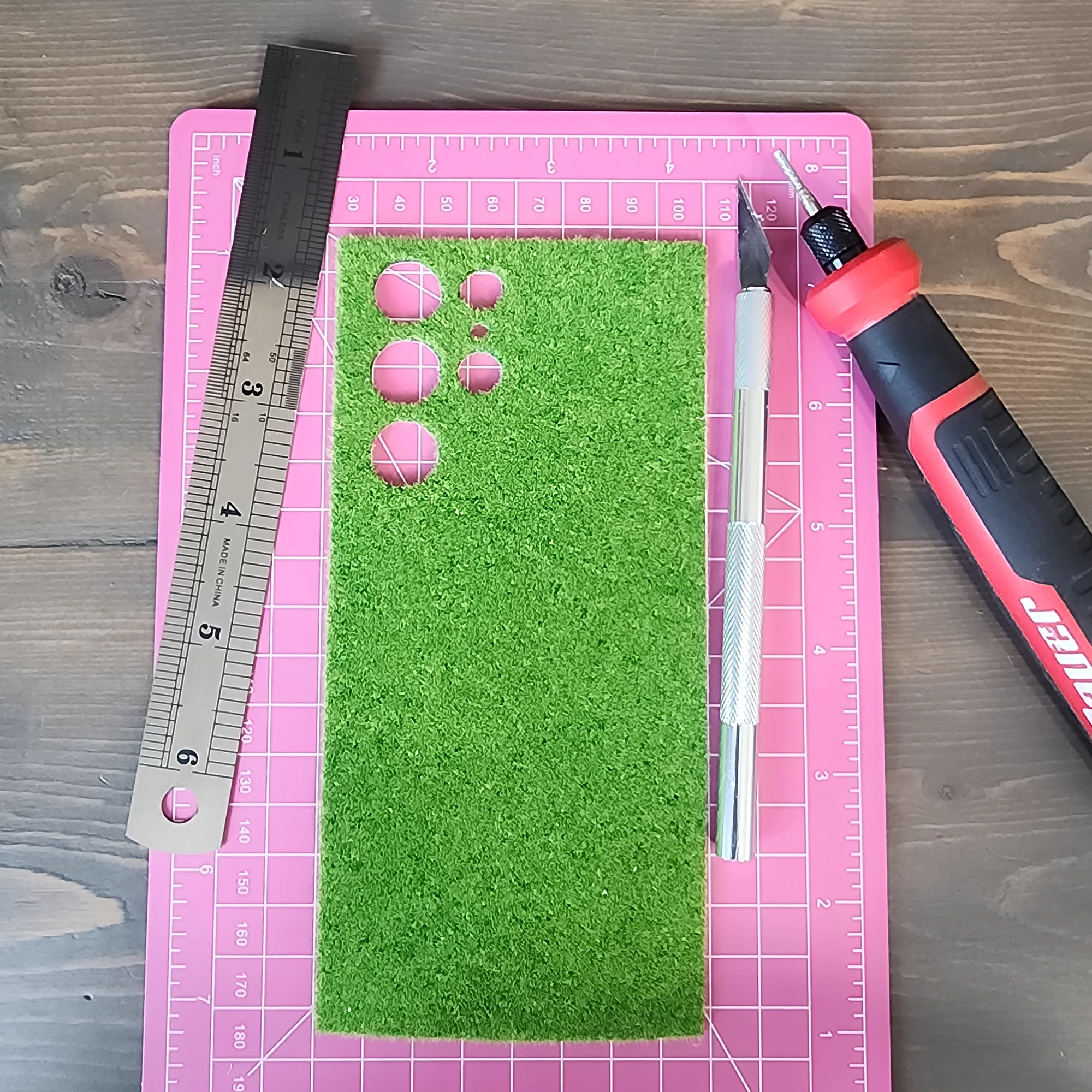 A small sheet of artificial grass being cut on a pink work mat with hand tools nearby.